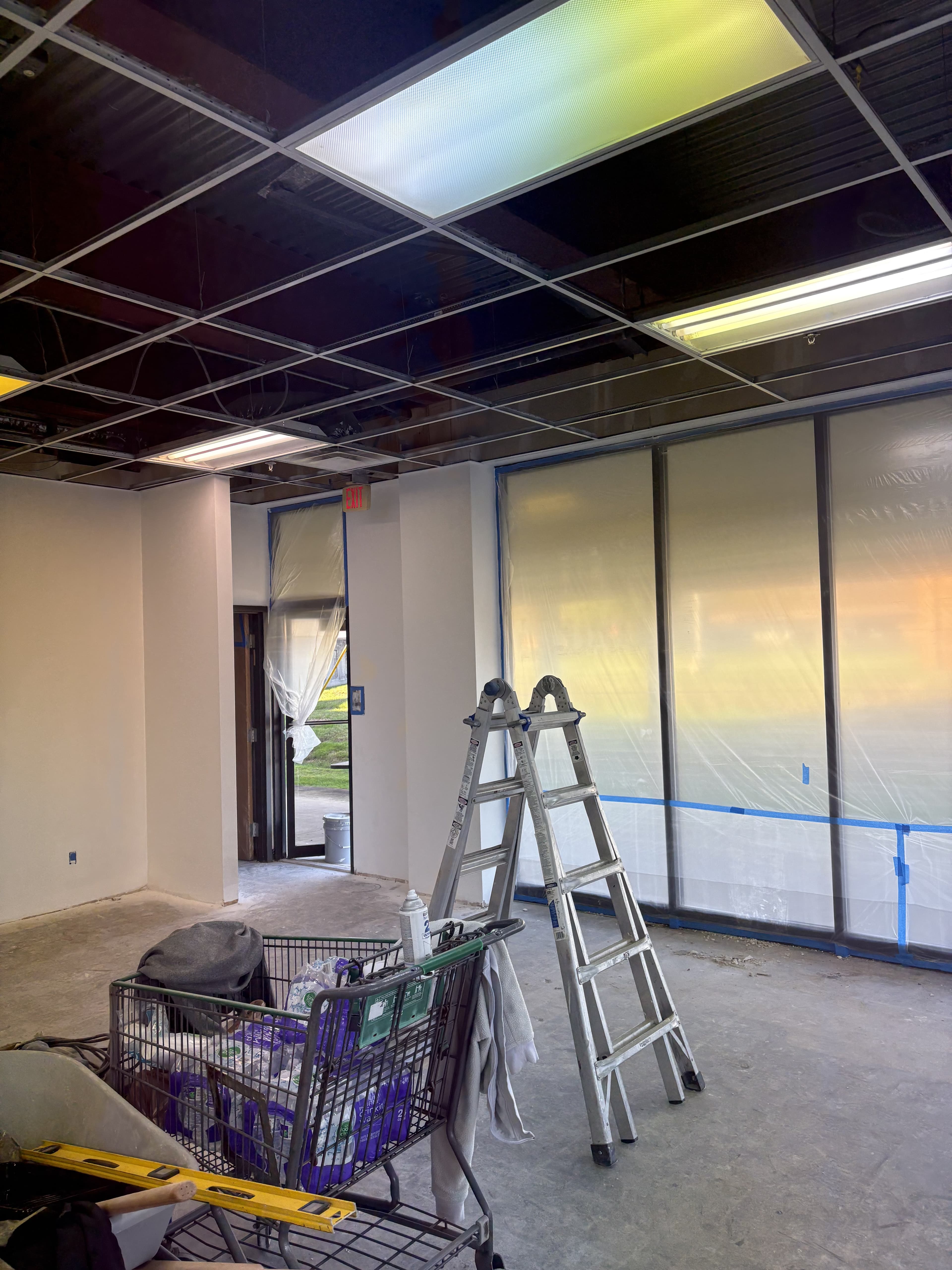 Commercial room with masked storefront windows, open ceiling grid, and active renovation work.